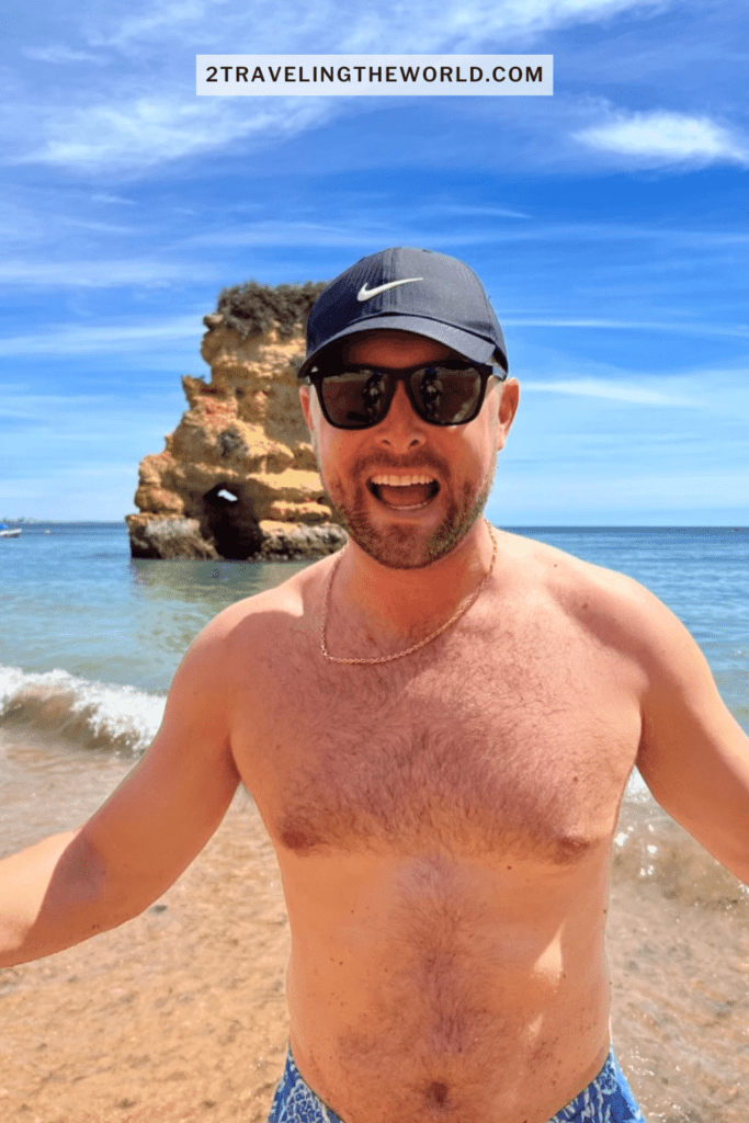 man standing on a beach in Southern Europe on one of the most stunning algarve beaches. He has a black Nike hat on and has Algarve rock formations coming out of the water behind him.