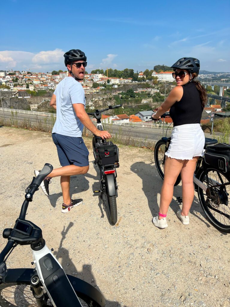 bike tour stop at luís i bridge porto landmark. man is standing by bike kicking his foot in the air. Girls is smiling back at camera holding on to her bike.