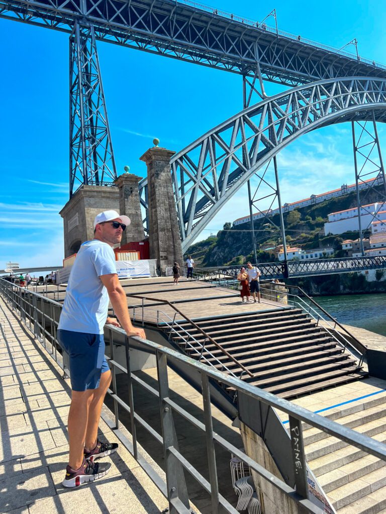 man standing next to the dom luis I Bridge over river douro lined by colorful buildings and hosues