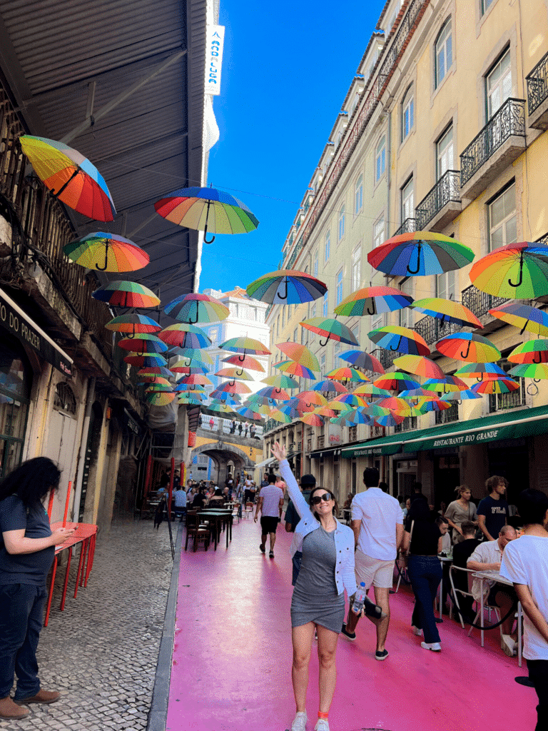 Pink Street in beautiful city of Lisbon. Girl has her hand up in the air. The street below her has bright pink paint. Their are rainbow color umbrellas hanging over the streets