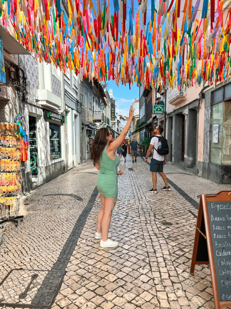 day trip to Aviero and Costa Nova with an organized tour. Shows girl in a green dress reaching up to touch colorful ribbons hanging across the buildings in Aviero Portugal