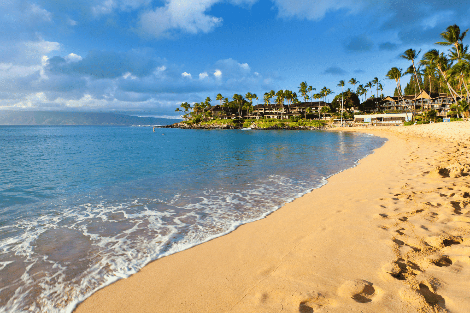 image of Napili white sand beach next to the Napili Kai Beach Resort with west maui mountains in the distance making it the perfect stay in maui