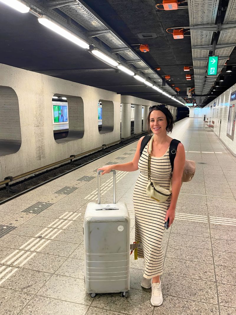 2TravelingTheWorld Team in the On Cloud 5 Slip ons standing next to a train station with her suitcase