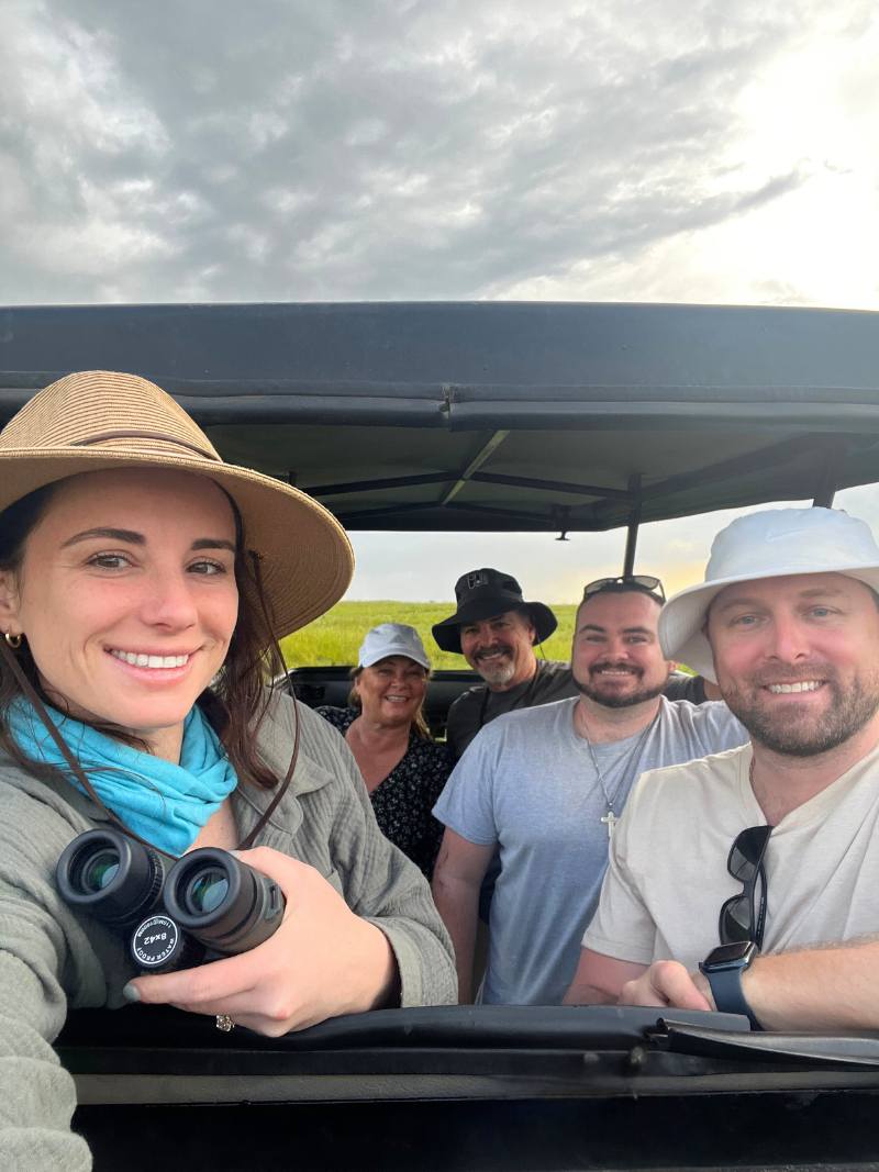 family selfie in an African safari game drive vehicle. Everyone is smiling at the camera. Woman has camera gear and binoculars in her hand